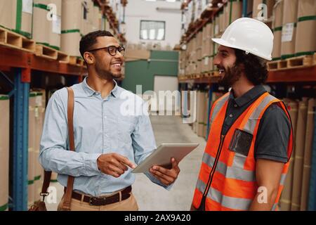 Warehouse manager wearing in formal clothing talking to loader in aisle between tall shelves with packed goods in modern factory