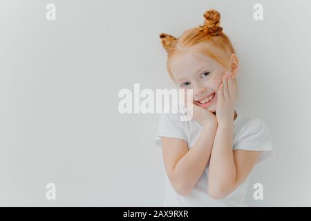 Shy pretty girl has two hair buns, touches cheeks gently, smiles happily, has freckles, hears something positive from parents, wears white t shirt, st Stock Photo