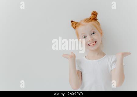 Playful red haired girl raises palms, has double buns, looks happily at camera, wears casual white t shirt, poses indoor, makes funny face, has toothy Stock Photo