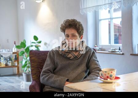 young man sits at the table at home with a scarf around his neck and has a thermometer in his mouth, it looks as if he has a cold Stock Photo