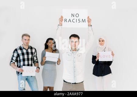 Handsome Caucasian young man protests with a poster, No racism concept, together with three multiethnic friends, African and Muslim women and Caucasia Stock Photo