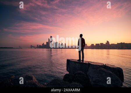 Young man standing on seashore and watching colorful sunrise. Sea and urban skyline Abu Dhabi, United Arab Emirates. Stock Photo