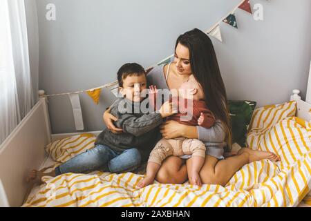 Young beautiful mother with her children on the bed, tightly hugs her son and little daughter, kisses her forehead, children laugh