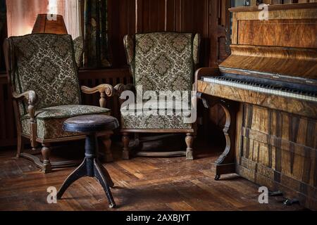 Vintage piano and a stool on a rustic and wooden floor with two old armchairs and a dirty window behind Stock Photo