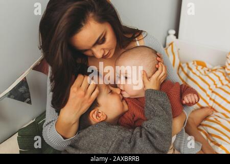 Young beautiful mother with her children on the bed, tightly hugs her son and little daughter, kisses her forehead, children laugh Stock Photo