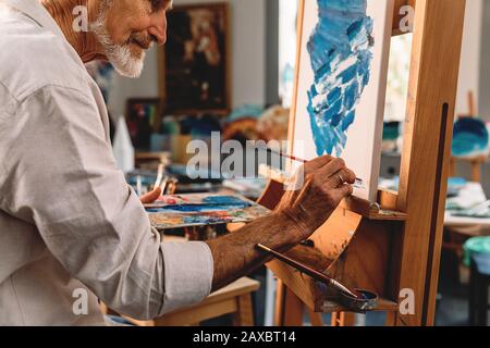 artist man is drawing on easel canvas, senior gray haired man in apron  enjoy the process of painting, in light room. side view Stock Photo - Alamy