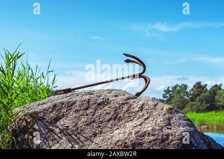 Old rusty boat anchor lying on a large stone against a blue sky. Stock Photo