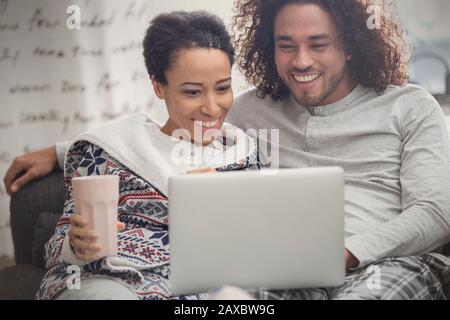 Happy couple in pajamas relaxing, using laptop on sofa Stock Photo