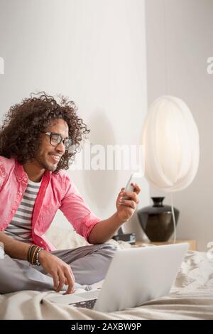 Smiling young man using smart phone at laptop on bed Stock Photo