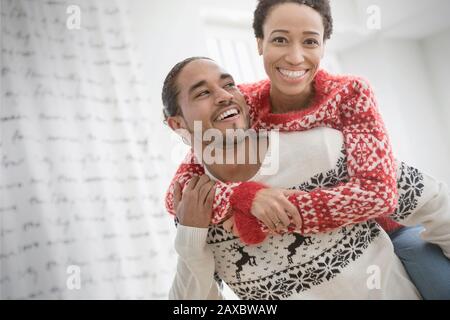 Portrait happy, playful couple in Christmas sweaters piggybacking Stock Photo