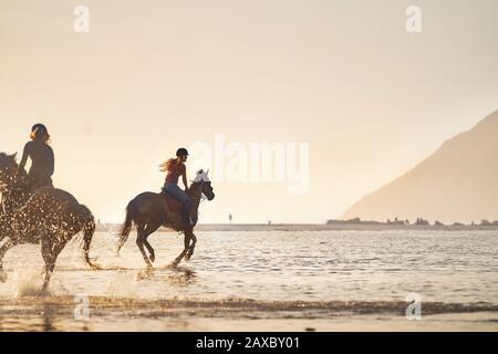 Young women horseback riding in ocean surf at sunset Stock Photo