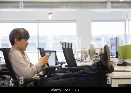 Businessman using smart phone with feet up on office desk Stock Photo