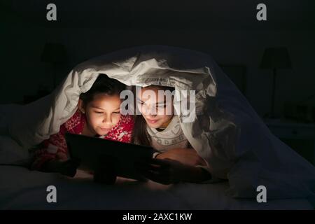 Sisters watching movie on digital tablet under blanket in dark bedroom Stock Photo