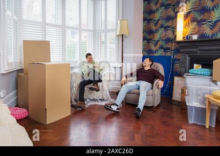 Men taking a break from moving house, relaxing and laughing in armchairs Stock Photo