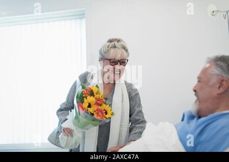 Happy senior woman bringing flower bouquet to husband recovering in hospital room Stock Photo