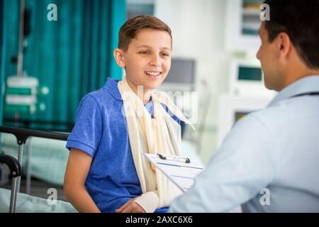 Male pediatrician talking to boy patient with arm in sling in clinic Stock Photo