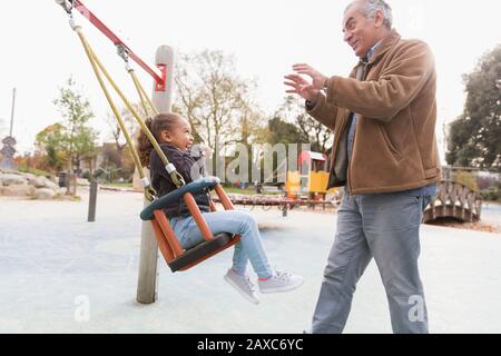 Grandfather pushing granddaughter on playground swing Stock Photo