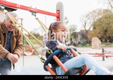 Playful grandfather pushing granddaughter on playground swing Stock Photo