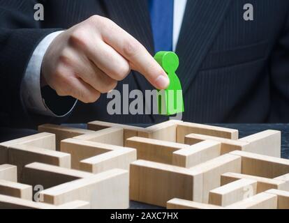 A male businessman holds a green figure over a maze. Make a path to success, provide support. Pass test, complete task. Professional skills developmen Stock Photo