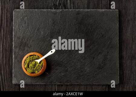 Matcha green tea powder in oak cup with white ceramic spoon with kitchen cutting board made of black slate. Stock Photo