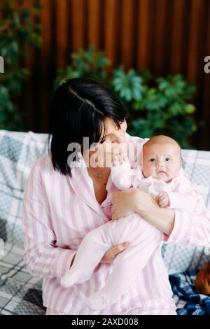 A mother in pajamas sits on a sofa and holds a baby in her arms. Stock Photo
