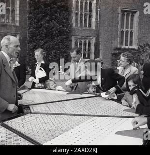 1950s, historical, in the grounds of a country house at a english summer garden party, well-dressed adults in the clothes of the day, with their children at a stall, playing a game of shove half penny or roll a penny, England, UK. A traditional game, often played in pubs and social clubs, it involves players pushing or rolling coins up a board. Stock Photo