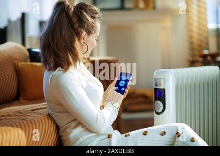 Stylish housewife in white sweater and skirt in the modern living room in sunny winter day sitting near couch and white electric oil radiator using sm Stock Photo