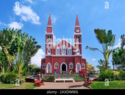 The all metal red and white catholic church in Grecia Costa Rica, sits between green palm trees and under a beautiful blue sky with white fluffy cloud