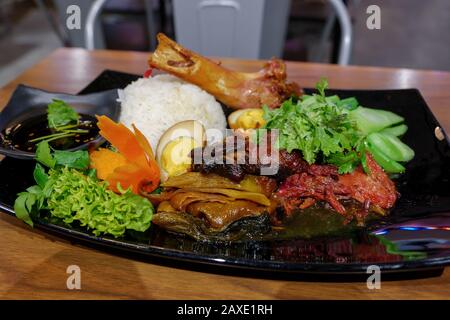 Stewed Pork leg and egg with various picked vegetable and steamed rice, on black dish and wooden table in restaurant. Stock Photo