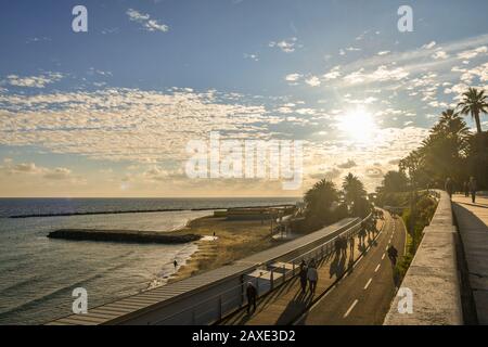 Backlit, scenic view of the waterfront with people and tourists walking and cycling on the pedestrian and bike path on the sea shore, Liguria, Italy Stock Photo
