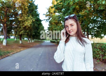 teenager girl 10-12 years old, holds smartphone in hand, listens voice message, calls mobile, summer on street park, beautiful thoughtful. Casual Stock Photo