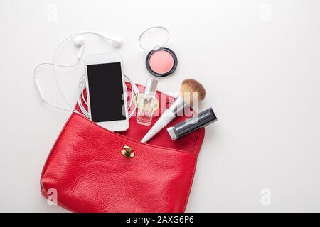 Red leather woman bag with lipstick, blush, perfume and smartphone on white background, top view Stock Photo