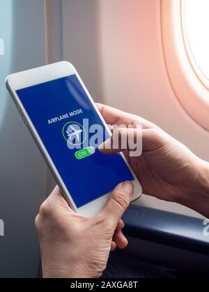 Flight mode concept. Hand holding white smartphone and turned on airplane mode on screen near the window on the airplane vertical style. Stock Photo
