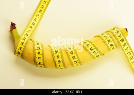 Centimeter ruler spinned around fresh fruit. Banana with yellow tape for measuring figure. Weight loss, diet and healthy lifestyle concept. Yellow tape around banana isolated on white background. Stock Photo