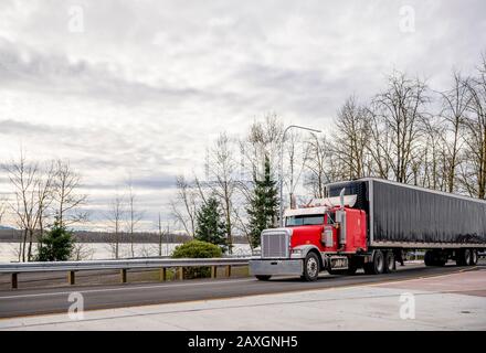 Big Rig red classic bonnet Semi Truck with Chrome Accessories and Vertical Exhaust Pipes transporting cargo in covered rubberized black semi trailer d Stock Photo