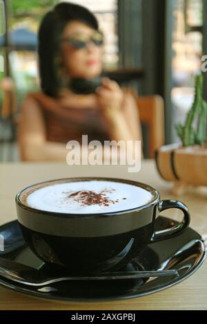 Closeup cappuccino in a black cup with blurry woman drinking coffee in the backdrop Stock Photo