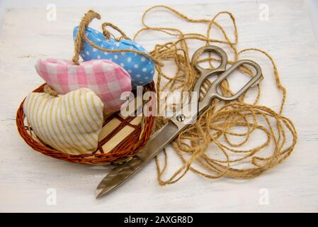 A basket filled with hearts sewn from scraps of fabric, next to large retro tailor's scissors, coarse thread and colorful buttons. Stock Photo