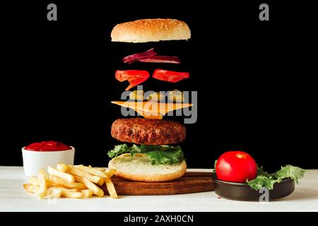 Floating beef burger. Tasty grilled meat with flying food ingredients on a black and white background with homemade ketchup and fries Stock Photo