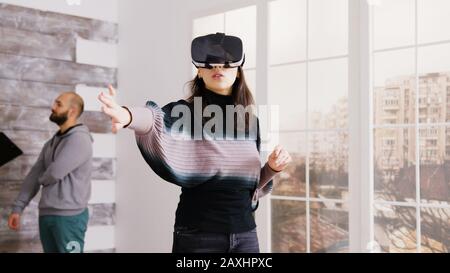 Female architect using virtual reality goggles in empty apartment and real estate agent talking with client in the background. Stock Photo