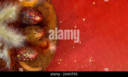 Top view of holiday card with gold red eggs, feathers with festive glitter confetti on a red background. Happy easter concept Stock Photo