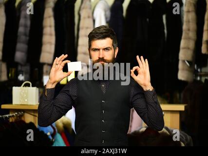 Finance and shopping concept. Businessman with empty paper shows ok sign. Guy with beard and business card. Man with serious face holds card on furry coats racks background, defocused Stock Photo