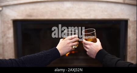 Glasses with whisky in male hands. Hands clinking whisky glasses on fireplace background. Male hands with drinks in glasses at home by fireplace. Men cheers with whisky in glasses near fireplace Stock Photo