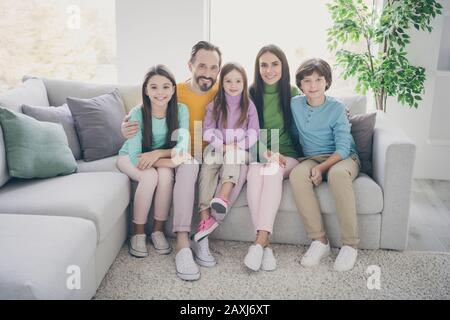 Full length photo of positive cheerful family three preteen kids schoolboy schoolgirl younger sister daddy mommy hug embrace sit couch chilling in Stock Photo