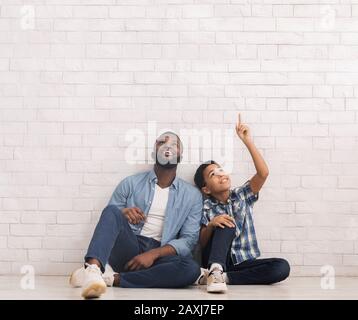 Father and son sitting on floor near white brick wall, pointing and looking upwards Stock Photo
