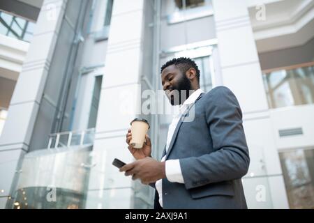 Below view of bearded Black businessman in jacket answering in messenger and drinking coffee on move Stock Photo