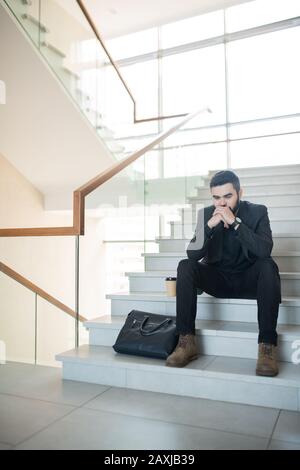 Stressed young businessman in black suit sitting on stairs with coffee cup and briefcase and leaning head on hands in puzzlement Stock Photo