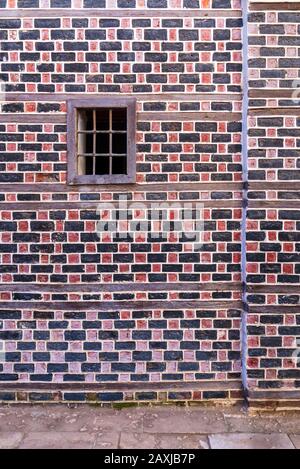 Wall with black and red bricks with white seam and small window with metal bars on cobblestone road Stock Photo