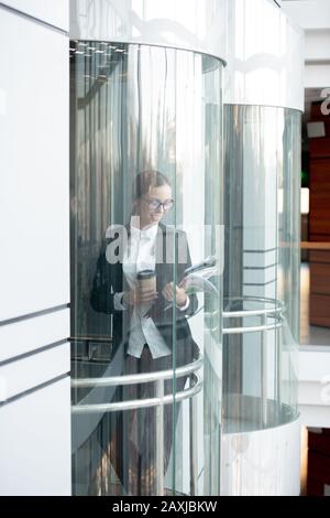 Smiling energetic young business lady in glasses drinking coffee and viewing papers in elevator of office building Stock Photo