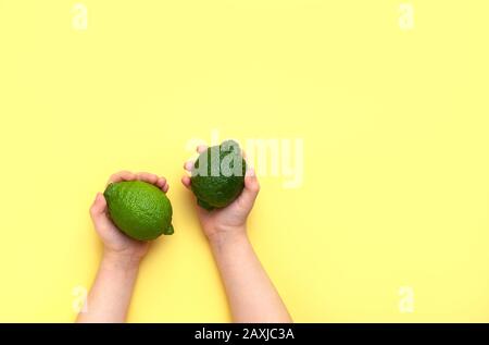 Children's hands hold two limes on a yellow background. Top view, copy space. Stock Photo