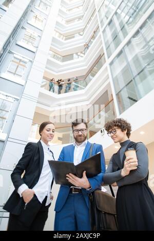 Below view of content young business team standing in office center and discussing data while analyzing papers Stock Photo